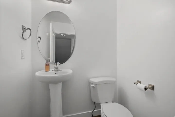 Pedestal sink featuring a chrome faucet, oval mirror with overhead lighting, and a white toilet with a brushed nickel toilet paper holder