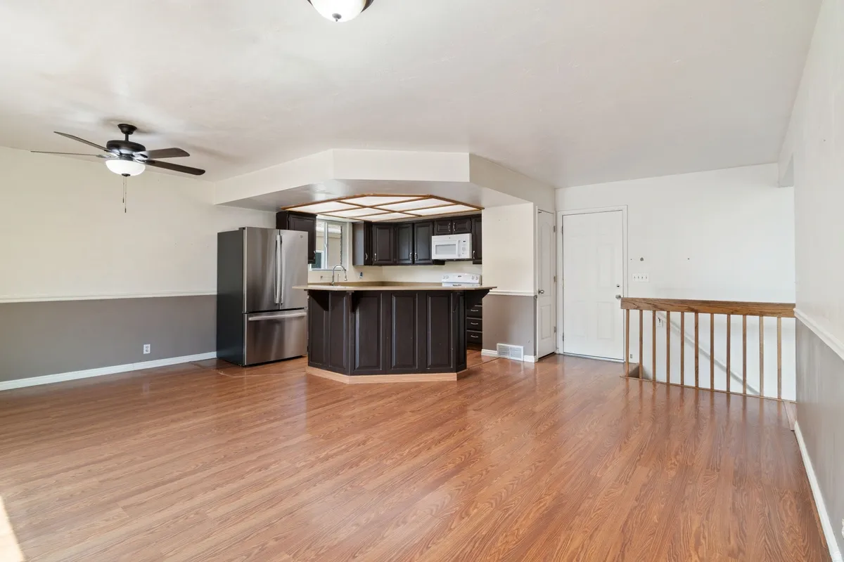 Open-concept living space featuring wood-finish flooring, a contemporary ceiling fan, and a kitchen area with dark cabinetry, stainless steel refrigerator, and white built-in microwave