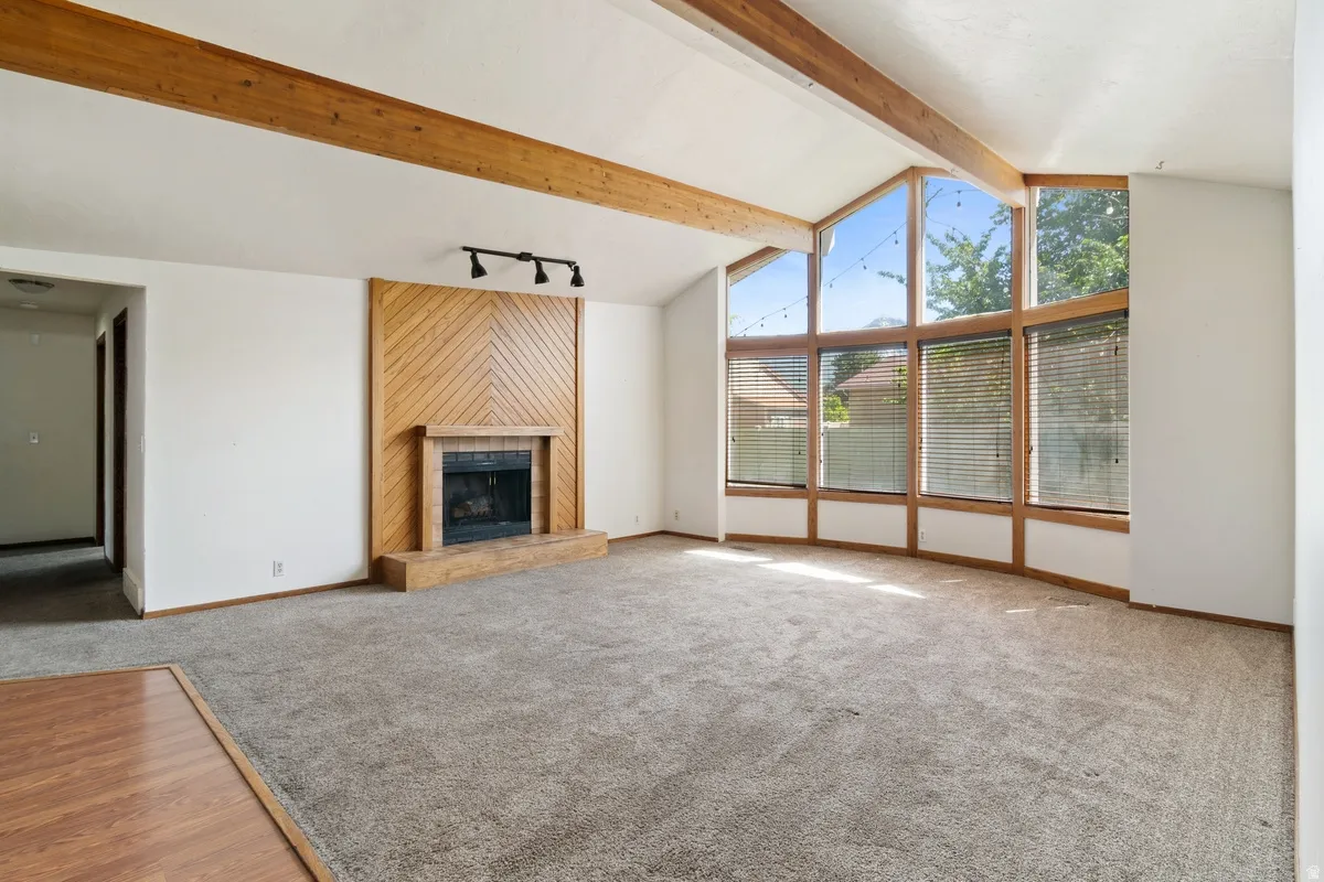 Vaulted ceiling with exposed wood beams, a prominent fireplace with a wood-paneled surround, and large picture windows featuring horizontal blinds