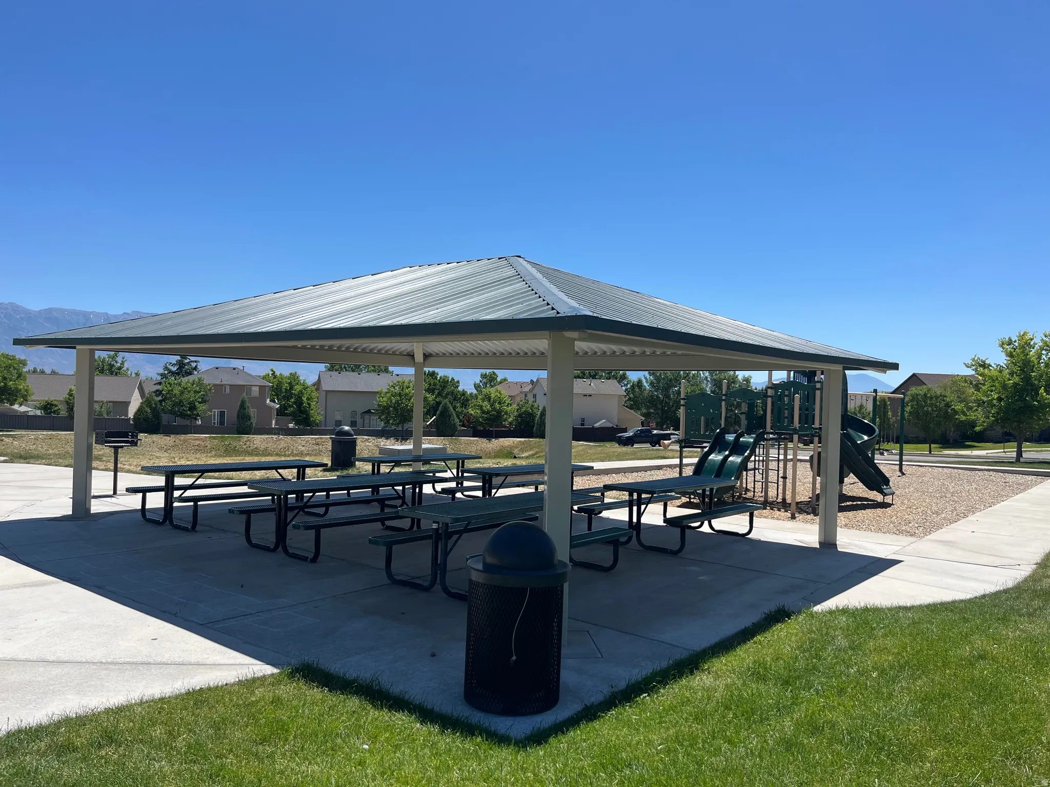 Covered outdoor seating area with multiple picnic tables, concrete flooring, an integrated barbecue grill, and a playground structure
