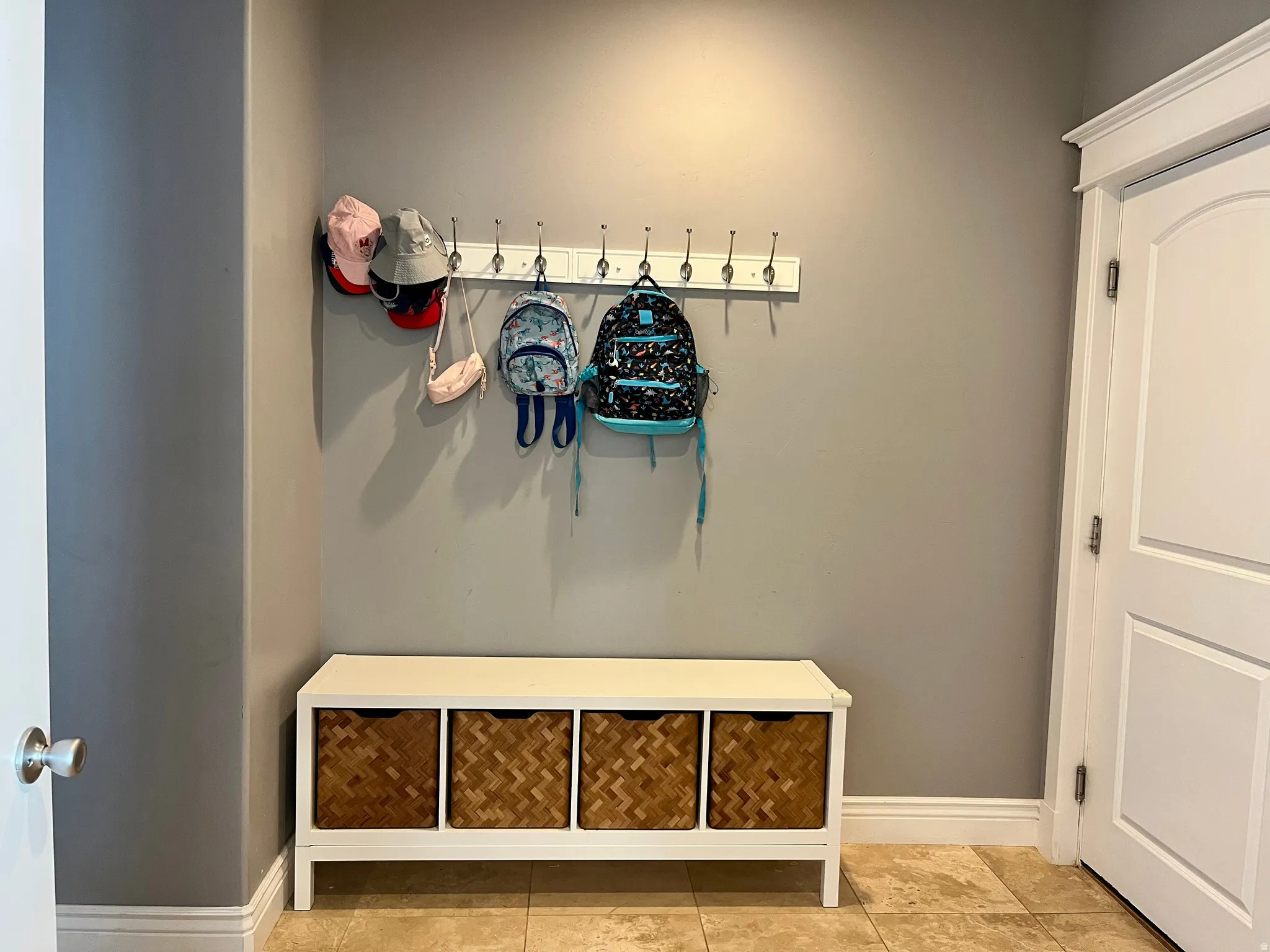 Built-in storage bench with four woven baskets, mounted coat rack with multiple hooks, neutral wall paint, and tile flooring