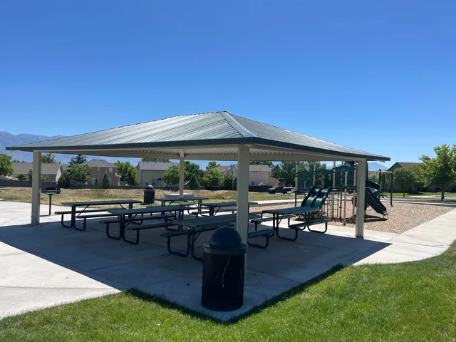 Covered outdoor seating area with multiple picnic tables, concrete flooring, an integrated barbecue grill, and a playground structure