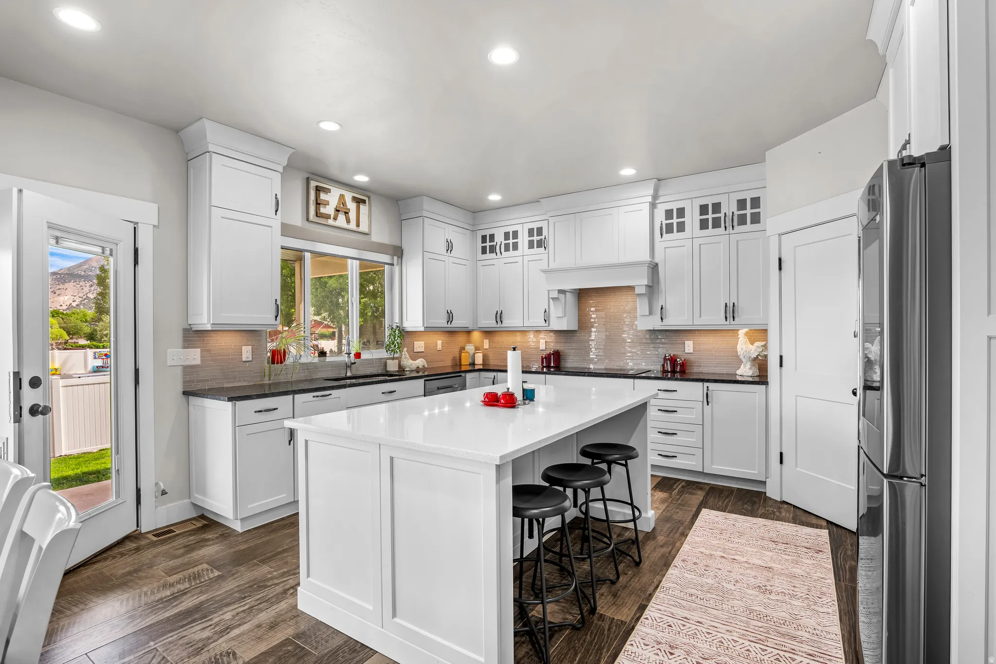 Contemporary kitchen featuring white shaker cabinetry, dark countertops, wood-finish flooring, a central island with bar seating, and a large window overlooking exterior scenery