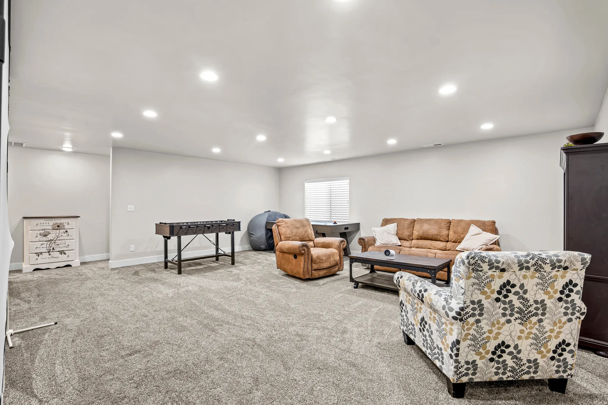Spacious finished room featuring light gray walls, plush gray carpeting, recessed ceiling lighting, a single window with blinds, and white trim