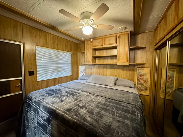 Bedroom featuring wood-finish wall paneling, a window with blinds, a ceiling fan with integrated lighting, and built-in wood cabinetry with open shelving