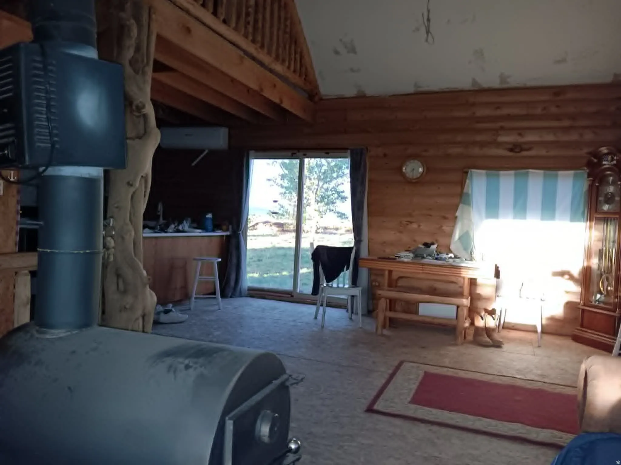 Spacious interior featuring natural wood walls, a wood-burning stove, exposed wood beam ceiling, an upper-level loft, and sliding glass door access to the outdoors
