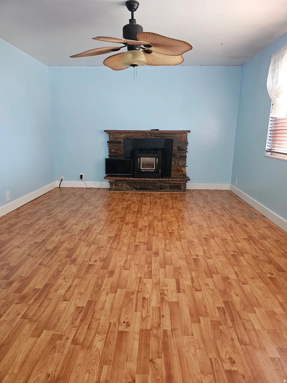 Spacious room featuring wood-finish flooring, a stone-clad fireplace with an insert, a ceiling fan with palm-leaf blades, and light blue wall finishes