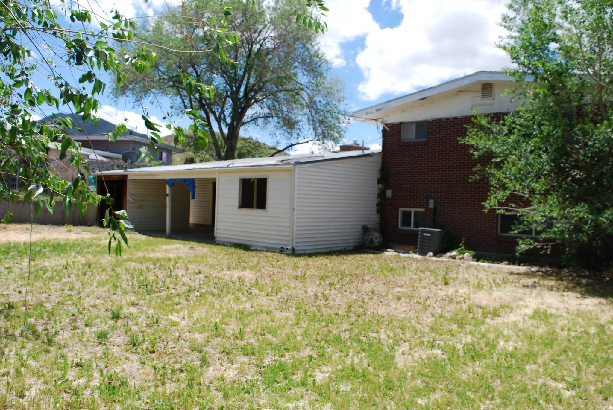 Expansive grass yard with a brick exterior home featuring multiple windows, an attached covered structure with additional windows, and mature trees