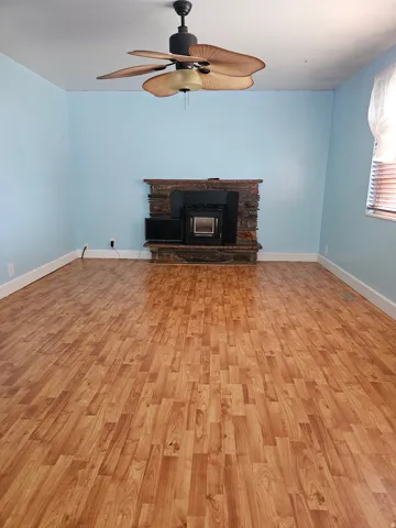 Spacious room featuring wood-finish flooring, a stone-clad fireplace with an insert, a ceiling fan with palm-leaf blades, and light blue wall finishes