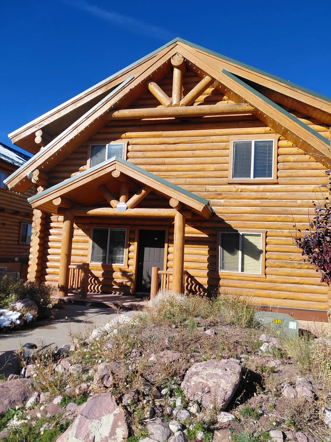 Log cabin exterior featuring a covered porch with log pillars, a gabled roofline, multiple windows with blinds, and natural rock landscaping