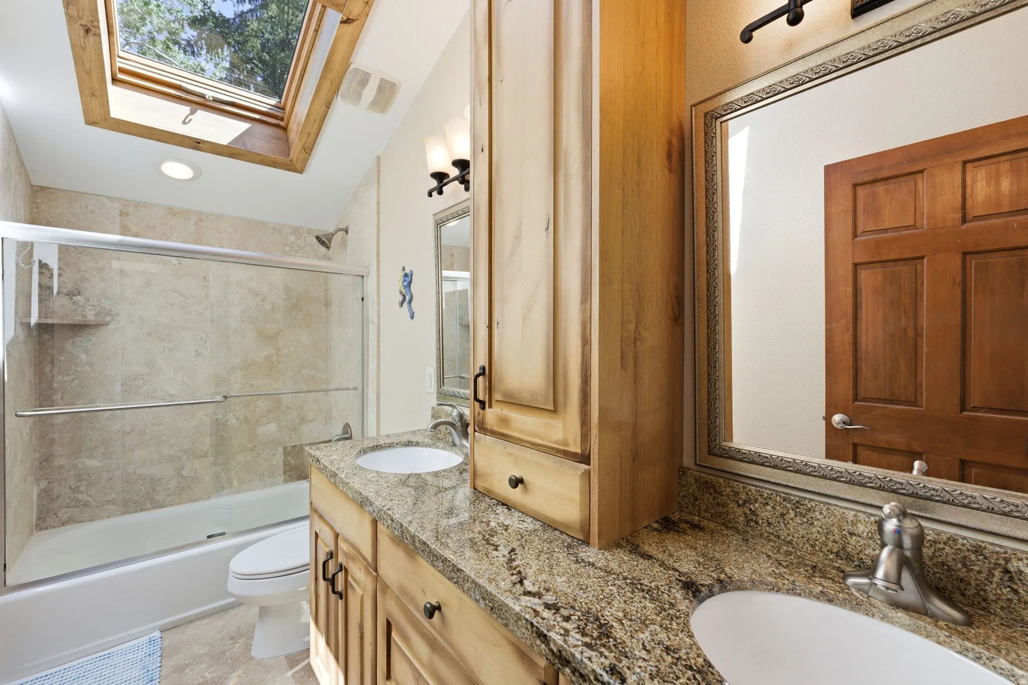 Bathroom featuring a skylight, dual vanity with undermount sinks, granite-finish countertops, wood cabinetry, and a shower-over-tub with sliding glass enclosure and tiled surround