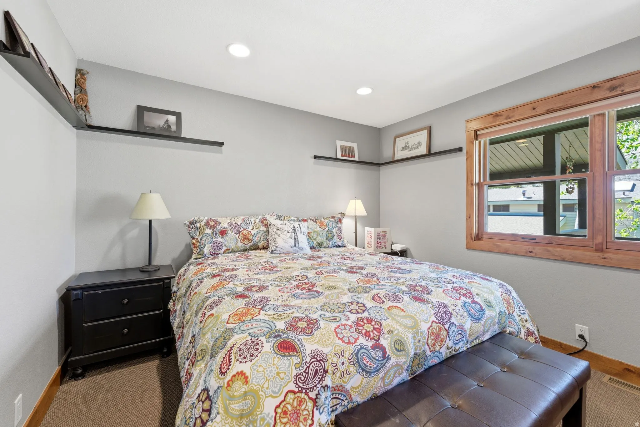 Recessed ceiling lighting illuminates a bedroom featuring light gray walls, wood-finish flooring, and a large window with natural wood trim