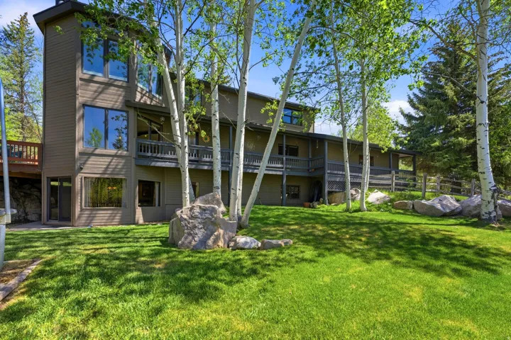 Multi-level home featuring extensive decking, a manicured lawn, mature trees, and natural rock landscaping