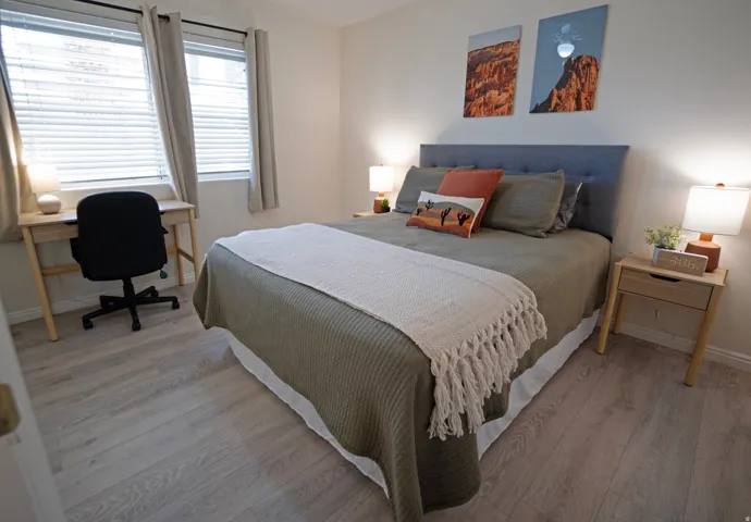 Bedroom featuring light wood-finish flooring, a tufted fabric headboard, two windows with blinds and curtains, and a built-in desk area