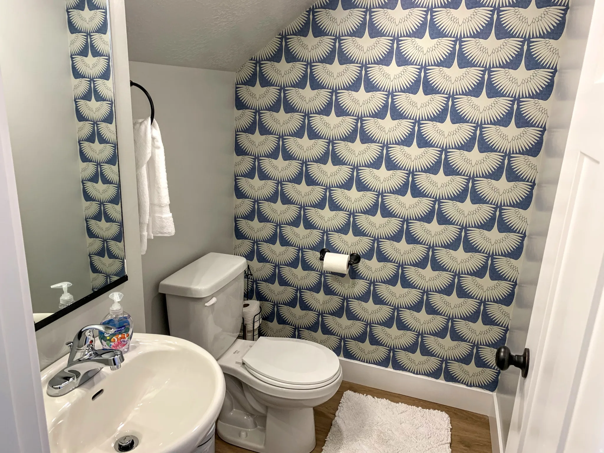 Powder room with patterned wallpaper, pedestal sink, chrome faucet, white baseboards, and wood-finish flooring