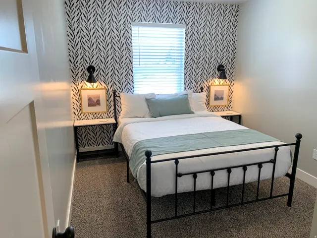 Bedroom featuring an accent wall with a striking black and white pattern, a window with horizontal blinds, built-in wall sconces, and neutral-toned carpeting