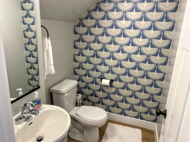 Powder room with patterned wallpaper, pedestal sink, chrome faucet, white baseboards, and wood-finish flooring