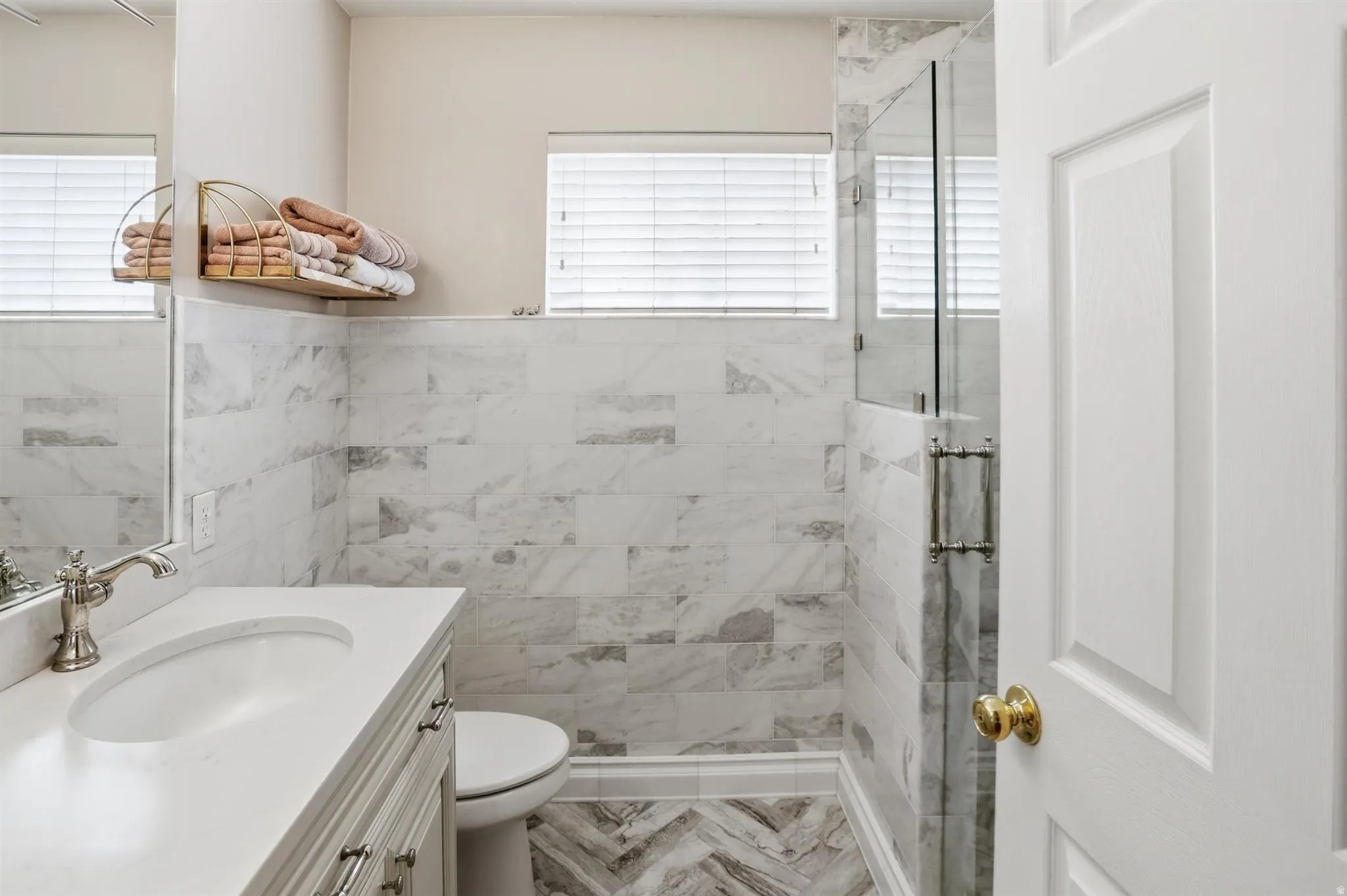 Modern bathroom featuring a glass-enclosed shower with marble-style tile surround, a single vanity with an undermount sink and light-colored countertop, and a chevron-patterned floor