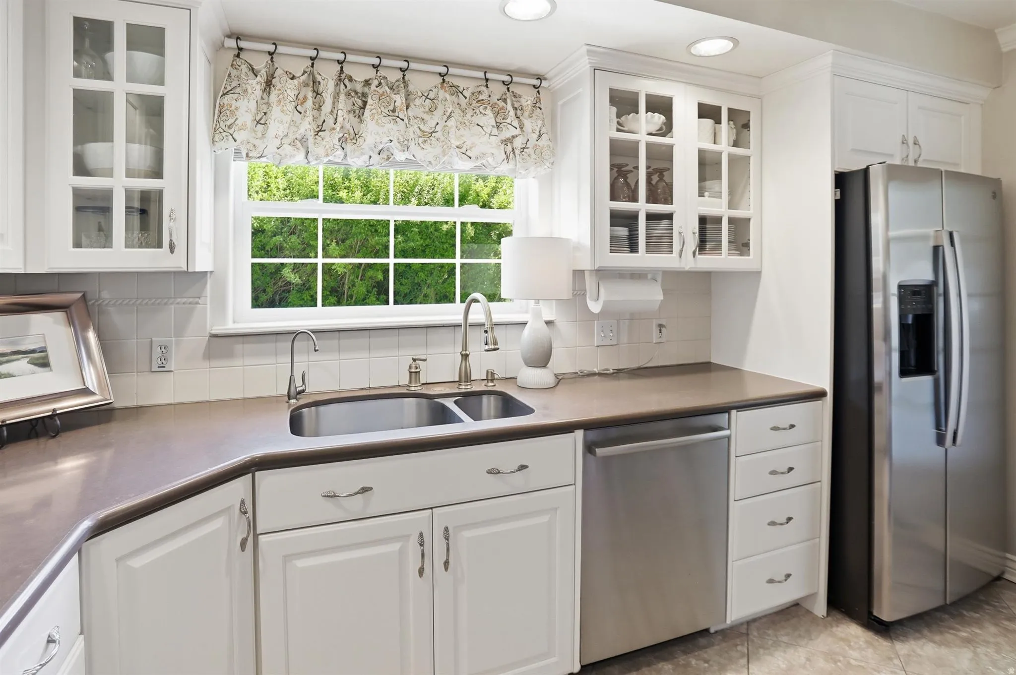 Kitchen featuring white cabinetry, a double basin stainless steel sink with a gooseneck faucet, light-colored countertops, and a full-size stainless steel refrigerator
