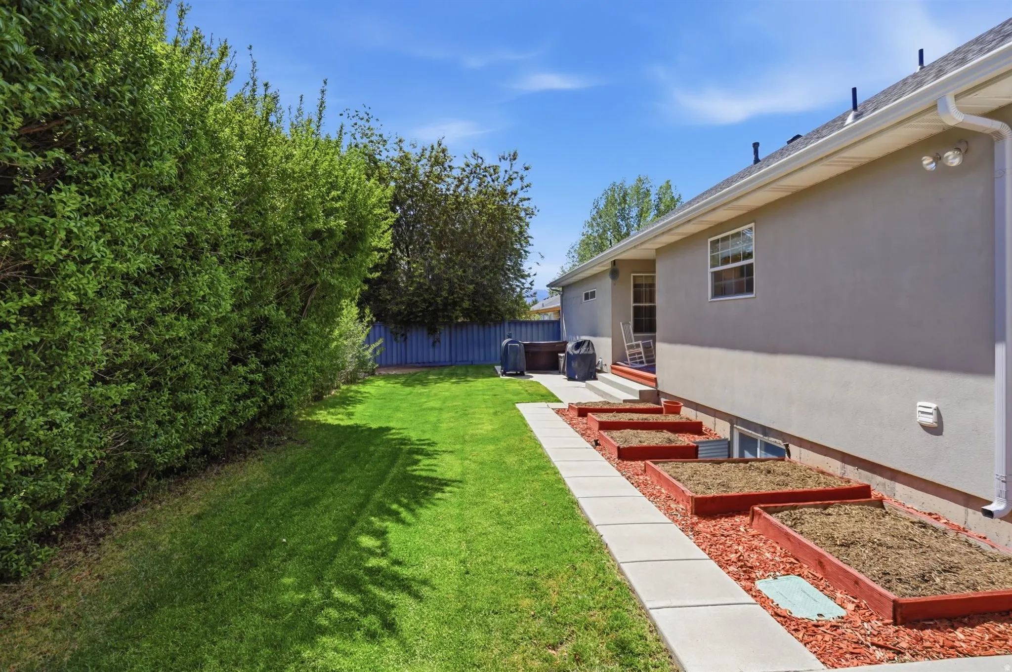 Expansive backyard featuring a green lawn, mature perimeter shrubbery, a concrete walkway, and tiered planter boxes with red mulch