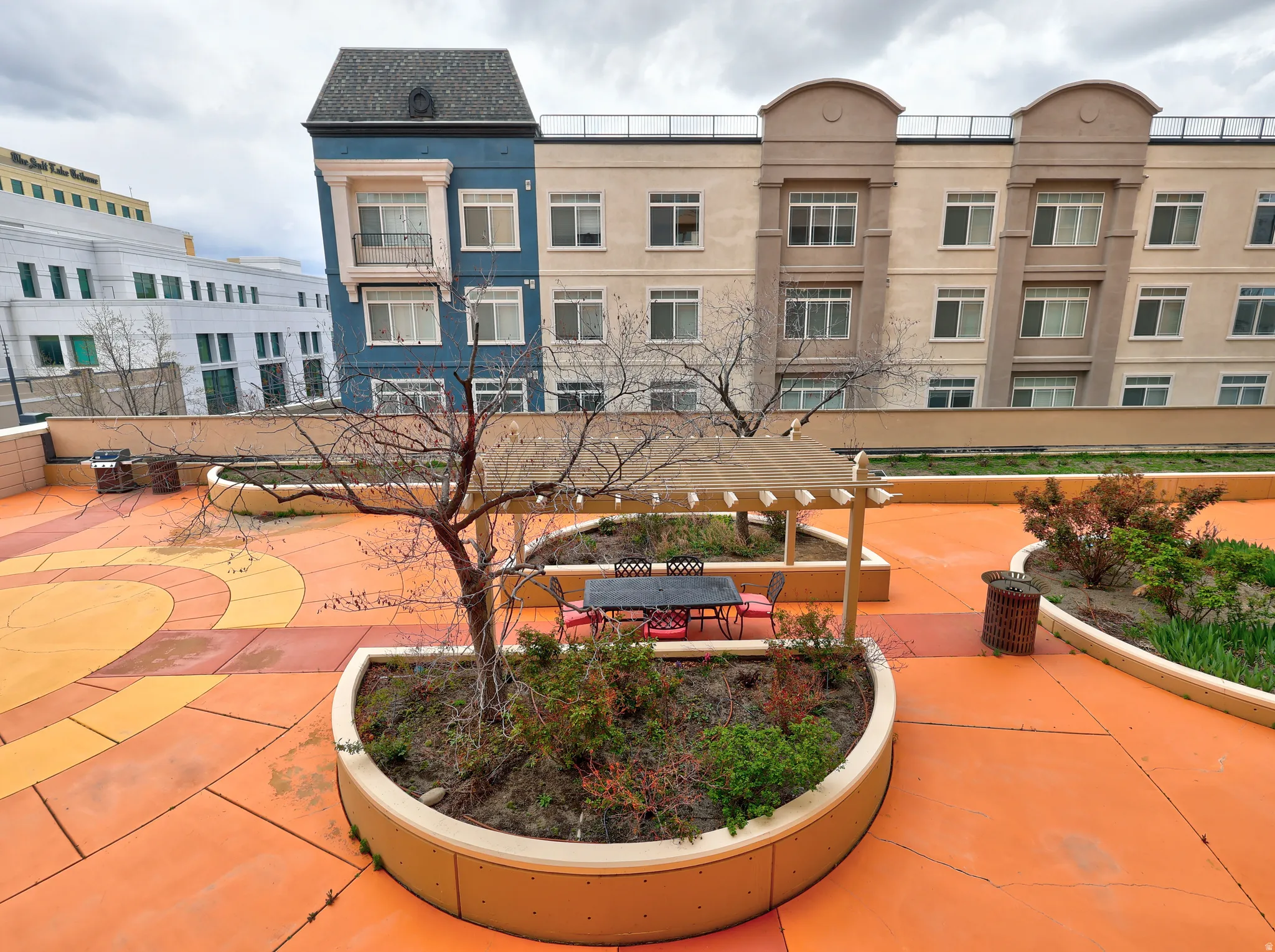 Rooftop patio featuring expansive orange and yellow patterned flooring, multiple built-in planter beds with mature trees and shrubs, and a shaded pergola
