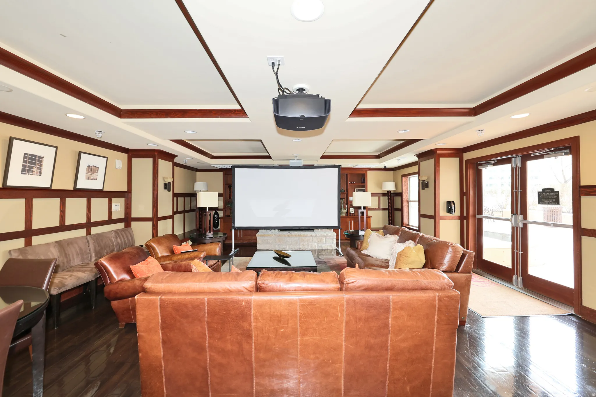 Spacious entertainment area featuring a ceiling-mounted projector, large screen, tray ceilings with recessed lighting, dark wood-finish flooring, and wainscoting