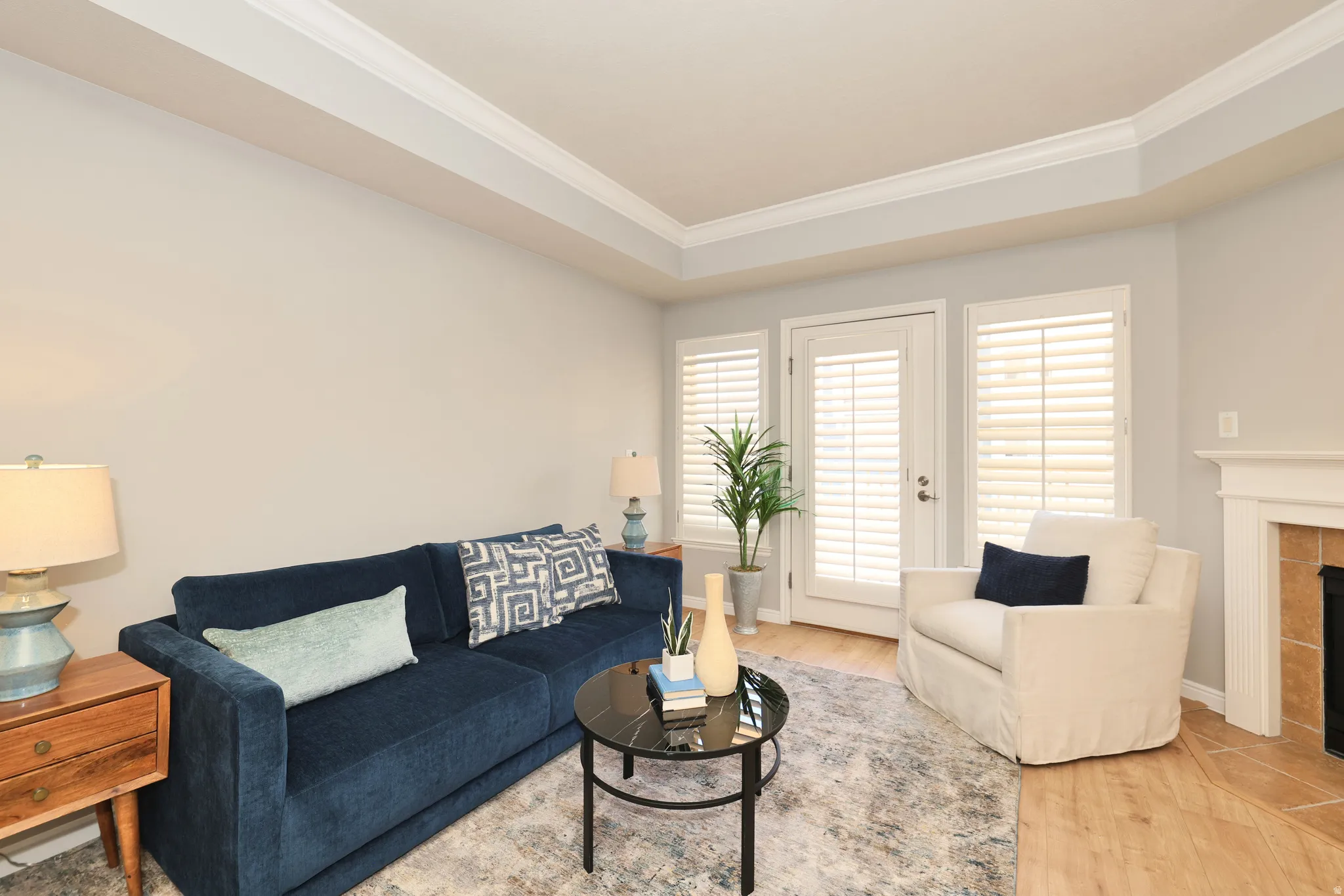 Living area featuring wood-finish flooring, a tray ceiling with crown molding, and a fireplace with a white mantel and tile surround