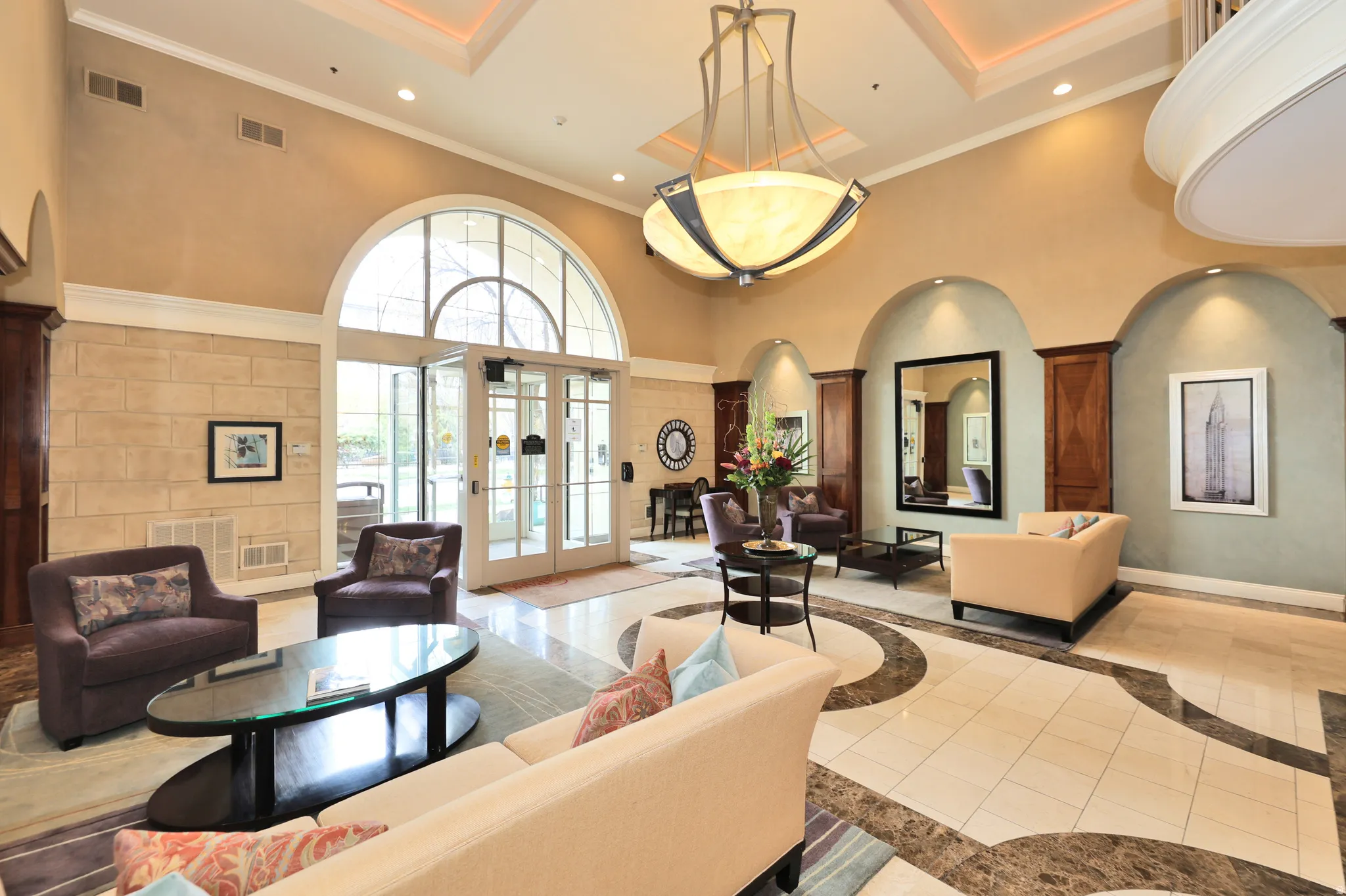 Grand foyer featuring a coffered ceiling with a large chandelier, arched doorway with a transom window, floor-to-ceiling stone-look wall treatment, and patterned tile flooring
