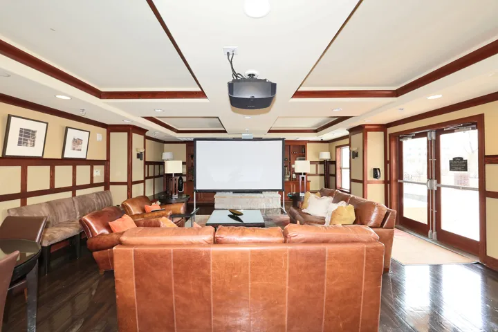 Spacious entertainment area featuring a ceiling-mounted projector, large screen, tray ceilings with recessed lighting, dark wood-finish flooring, and wainscoting