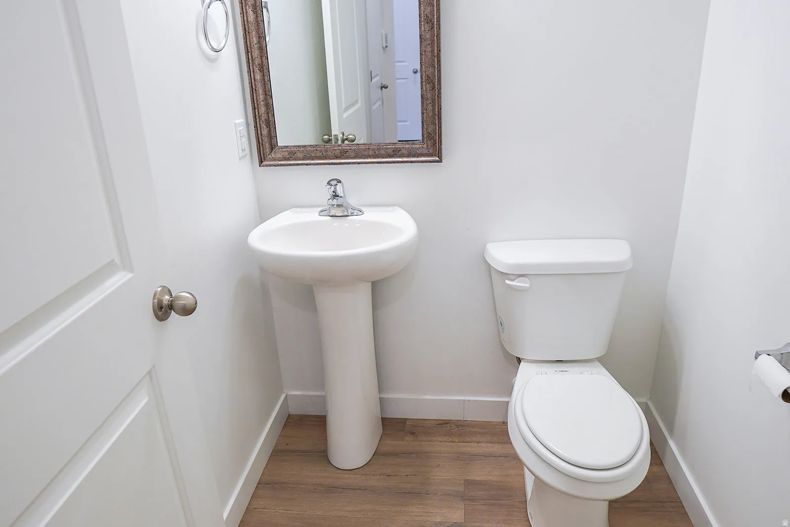 Pedestal sink with chrome faucet, white toilet, wood-finish flooring, and a framed mirror