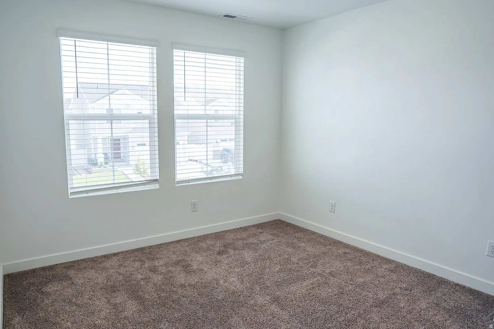 Bright interior room featuring a neutral color palette, two large windows, white window blinds, white baseboards, and brown textured carpeting