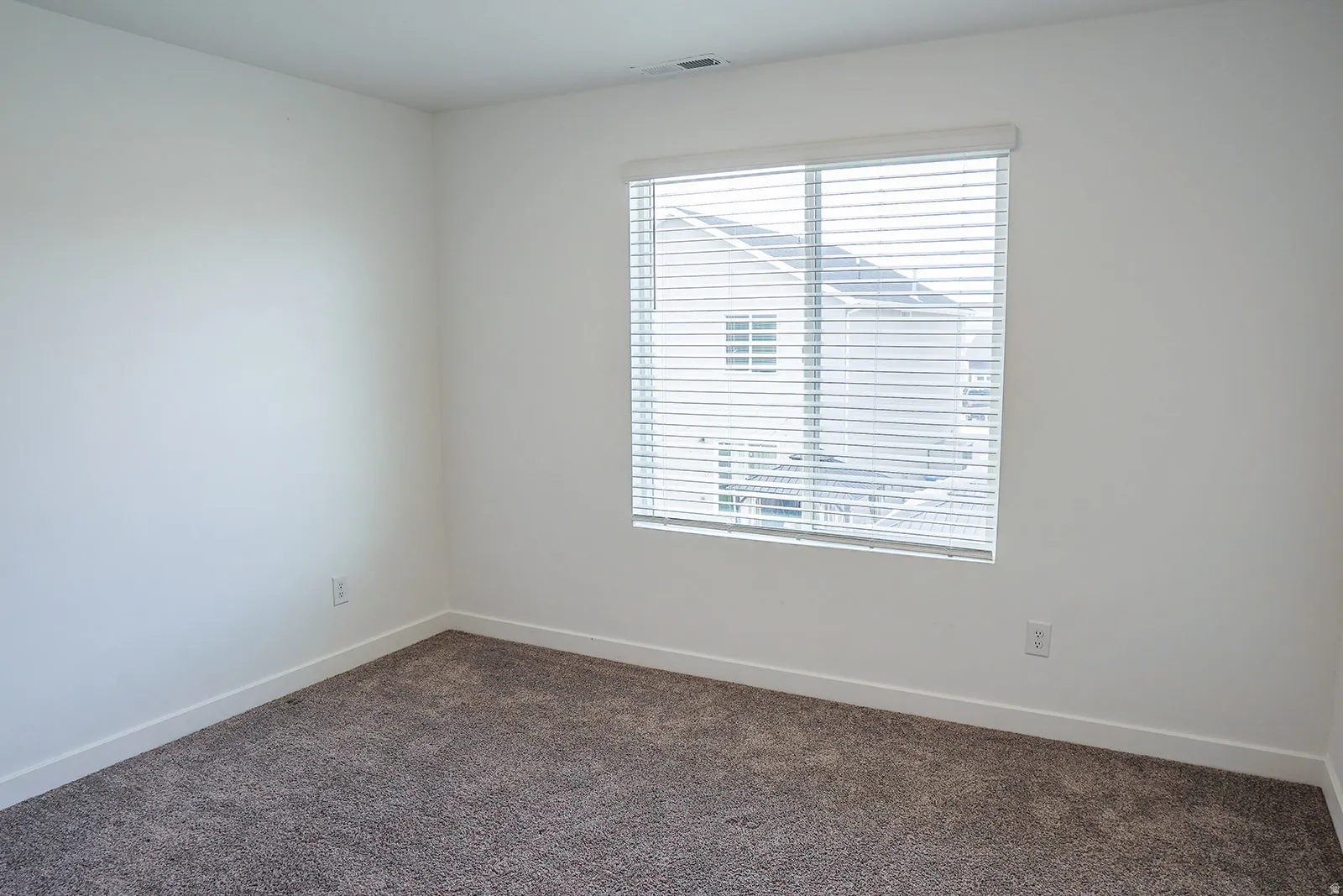 Neutral-toned room featuring a single window with blinds, light-colored walls, and dark-toned carpet