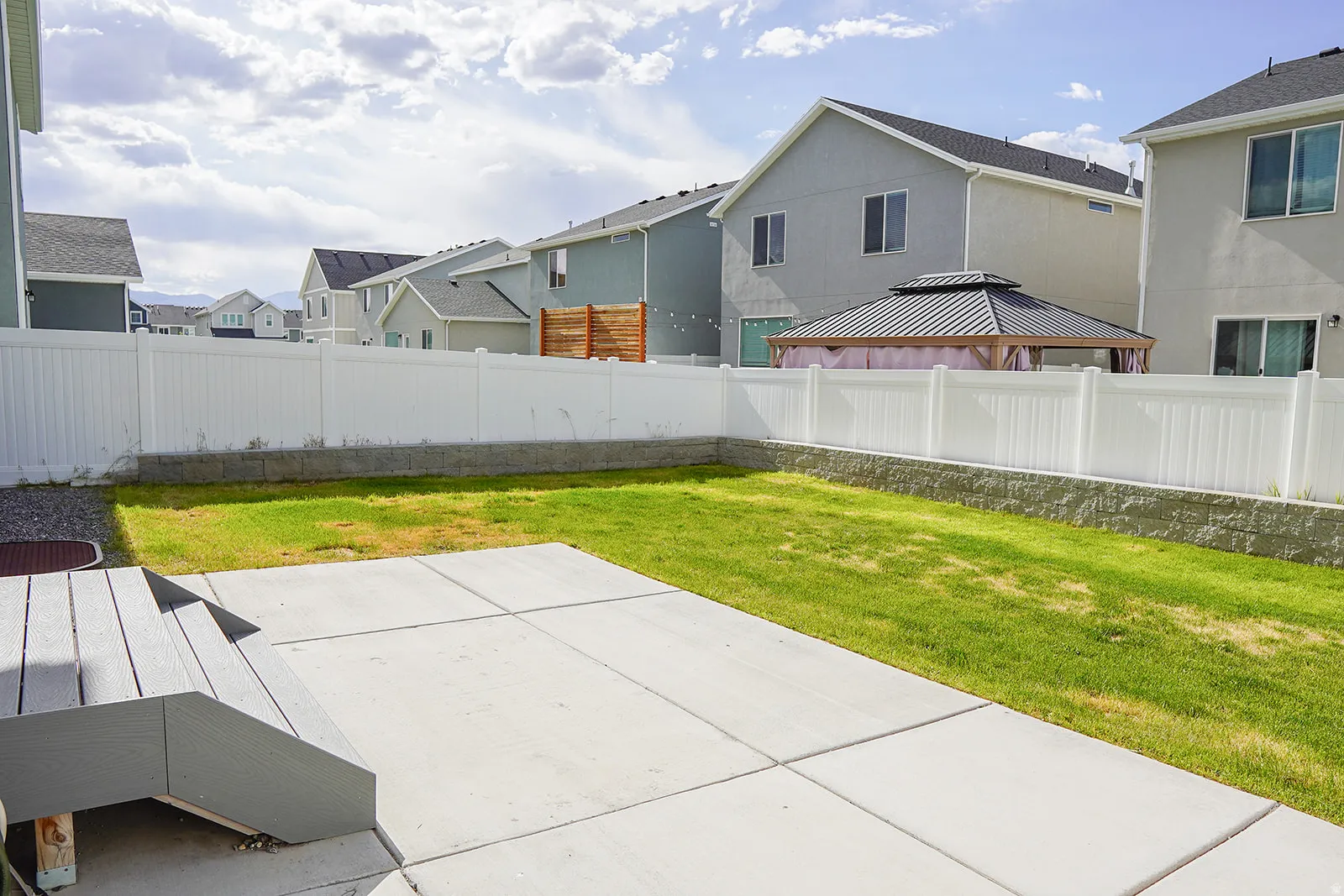 Expansive backyard featuring a concrete patio, composite deck steps, and a white vinyl privacy fence atop a stone retaining wall