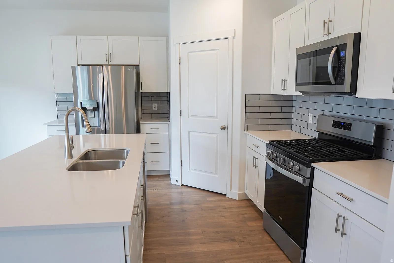 Contemporary kitchen featuring a large center island with double basin sink, white cabinetry with silver hardware, light-toned countertops, a stainless steel gas range, and a gray subway tile backsplash