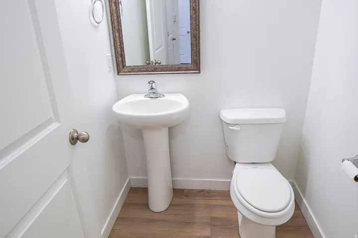 Pedestal sink with chrome faucet, white toilet, wood-finish flooring, and a framed mirror