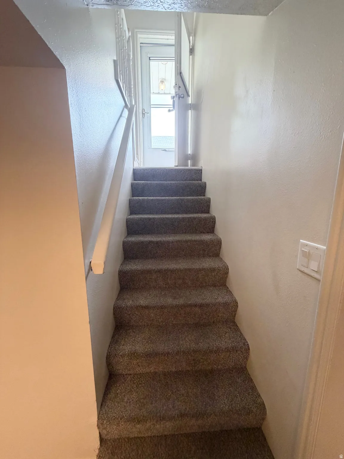 Carpeted staircase featuring a white handrail, white walls, and a white interior door at the top