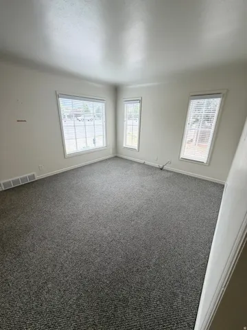 Spacious room featuring neutral carpeting, multiple windows with blinds, a baseboard heating vent, and light-toned walls