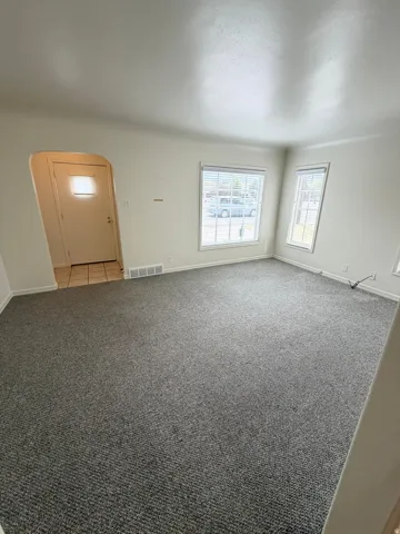 Spacious living area featuring neutral-toned carpeting, light-colored walls, multiple windows with blinds, a tiled entryway, and an arched doorway
