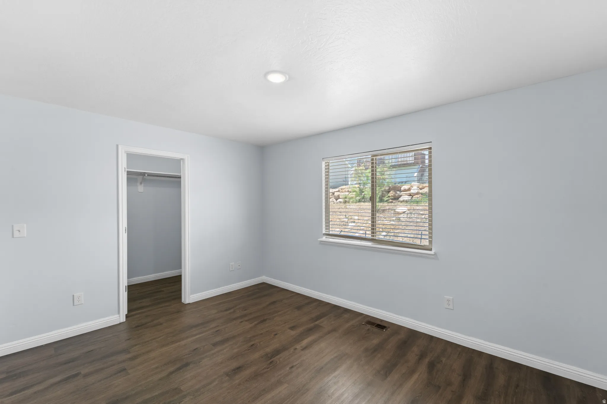 Spacious room featuring wood-finish flooring, light blue wall paint, a window with blinds, a recessed ceiling light, and white baseboards