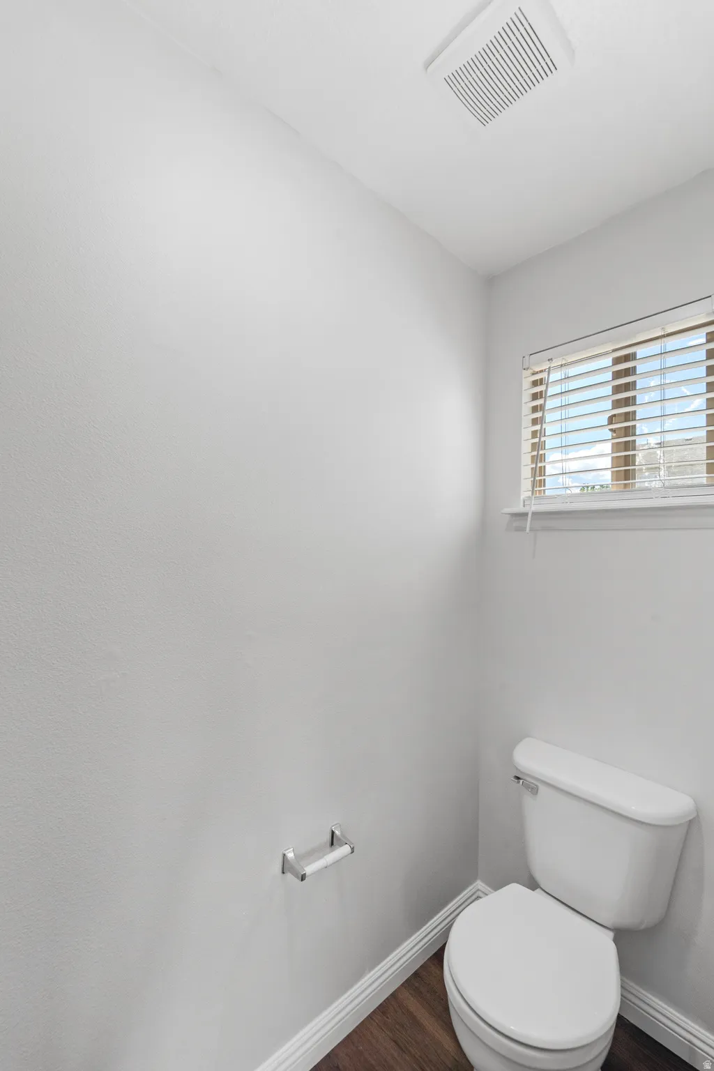 Powder room featuring a white toilet, wood-finish flooring, white baseboards, and a window with horizontal blinds