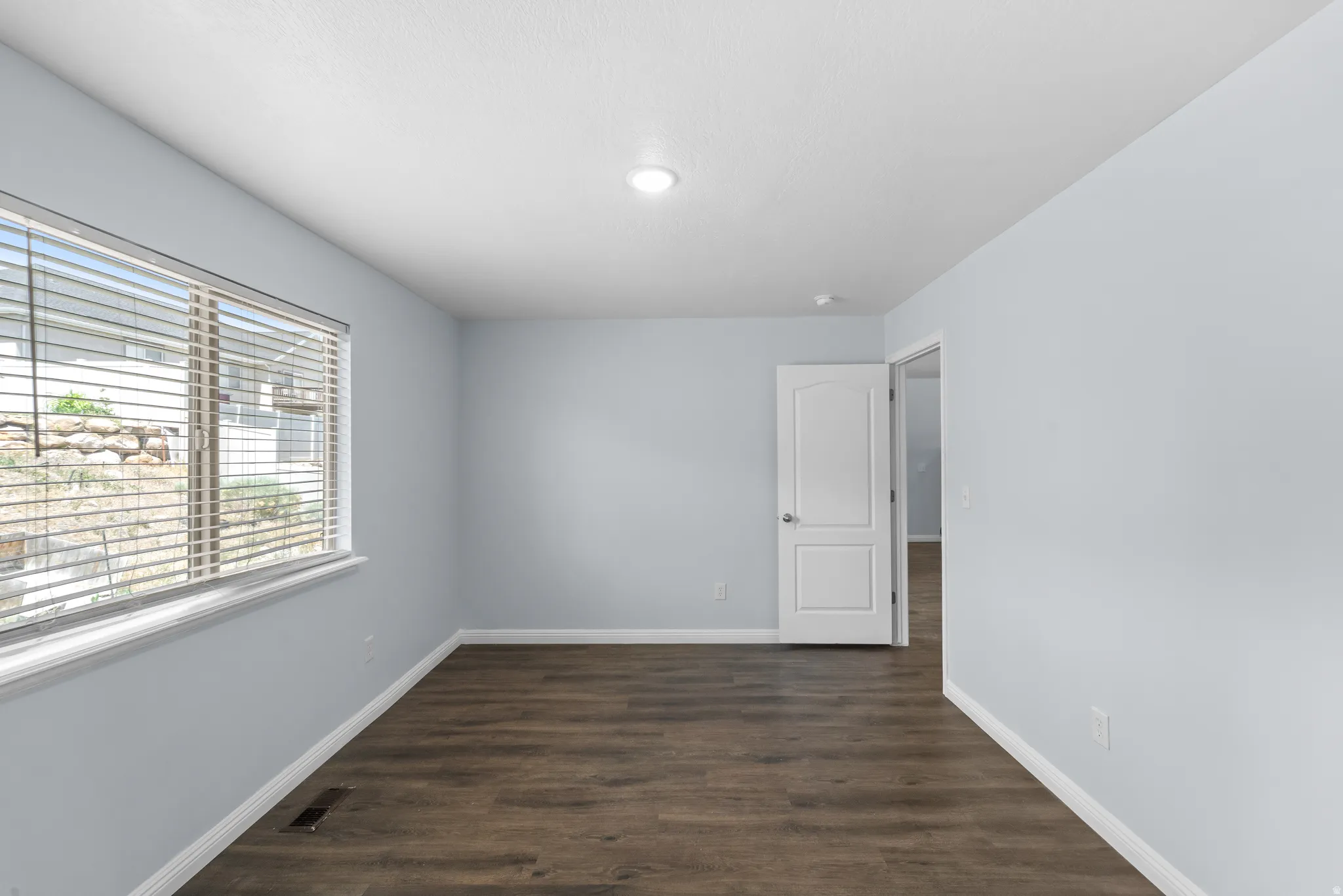 Spacious room featuring wood-finish flooring, light blue wall tones, and a large window with horizontal blinds