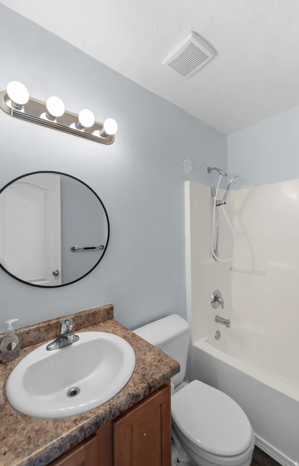 Bathroom featuring a white oval sink, a wood-finish vanity with a stone-finish countertop, a circular mirror, and a full-size bathtub with an integrated shower