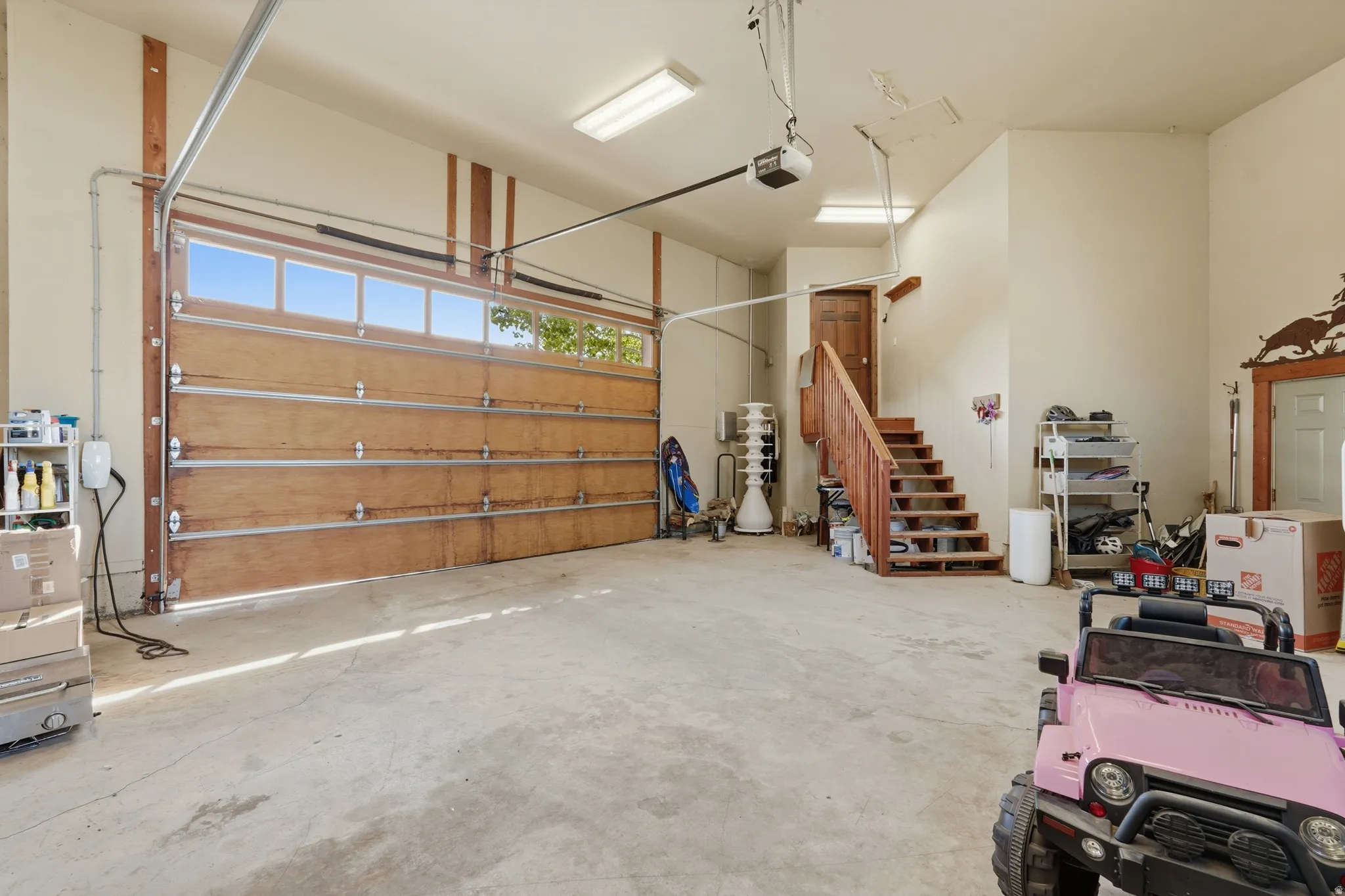 Spacious garage featuring a light wood-paneled garage door with upper window inserts, concrete flooring, wooden staircase with railing, recessed overhead lighting, and white painted walls