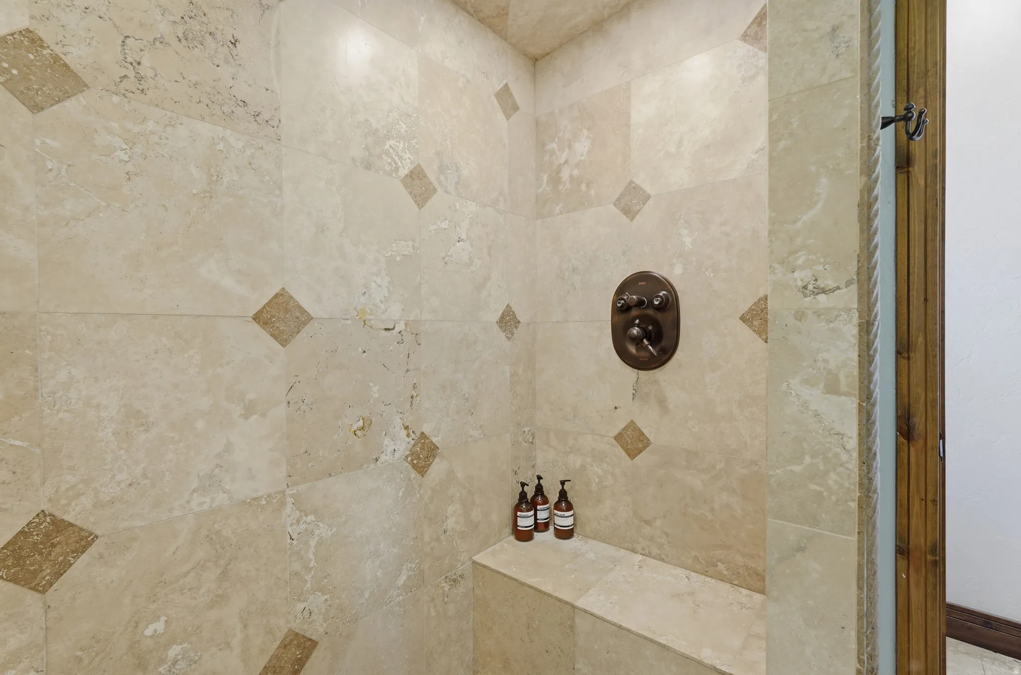 Shower with neutral-toned natural stone tilework, accented by diamond-patterned darker tiles, featuring a built-in bench and an oil-rubbed bronze shower control