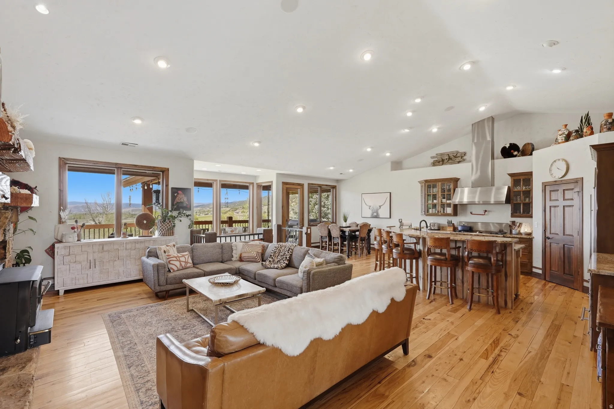 Open-concept living space featuring vaulted ceilings, wood-finish flooring, a stone fireplace, and an expansive window line
