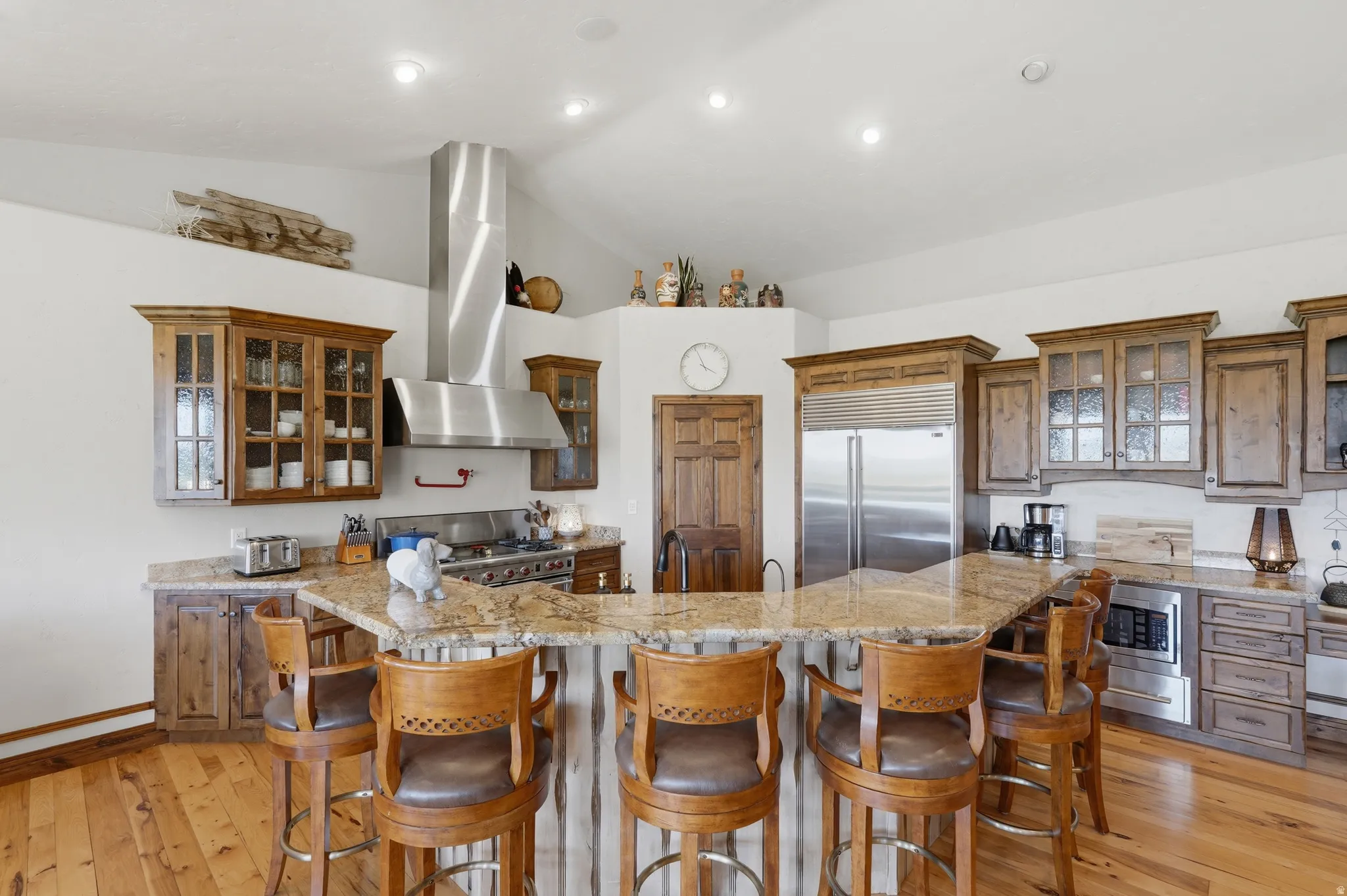Kitchen featuring a large granite island with bar seating, stainless steel range hood, built-in stainless steel refrigerator, wood-finish flooring, and ample cabinetry with glass-front accents