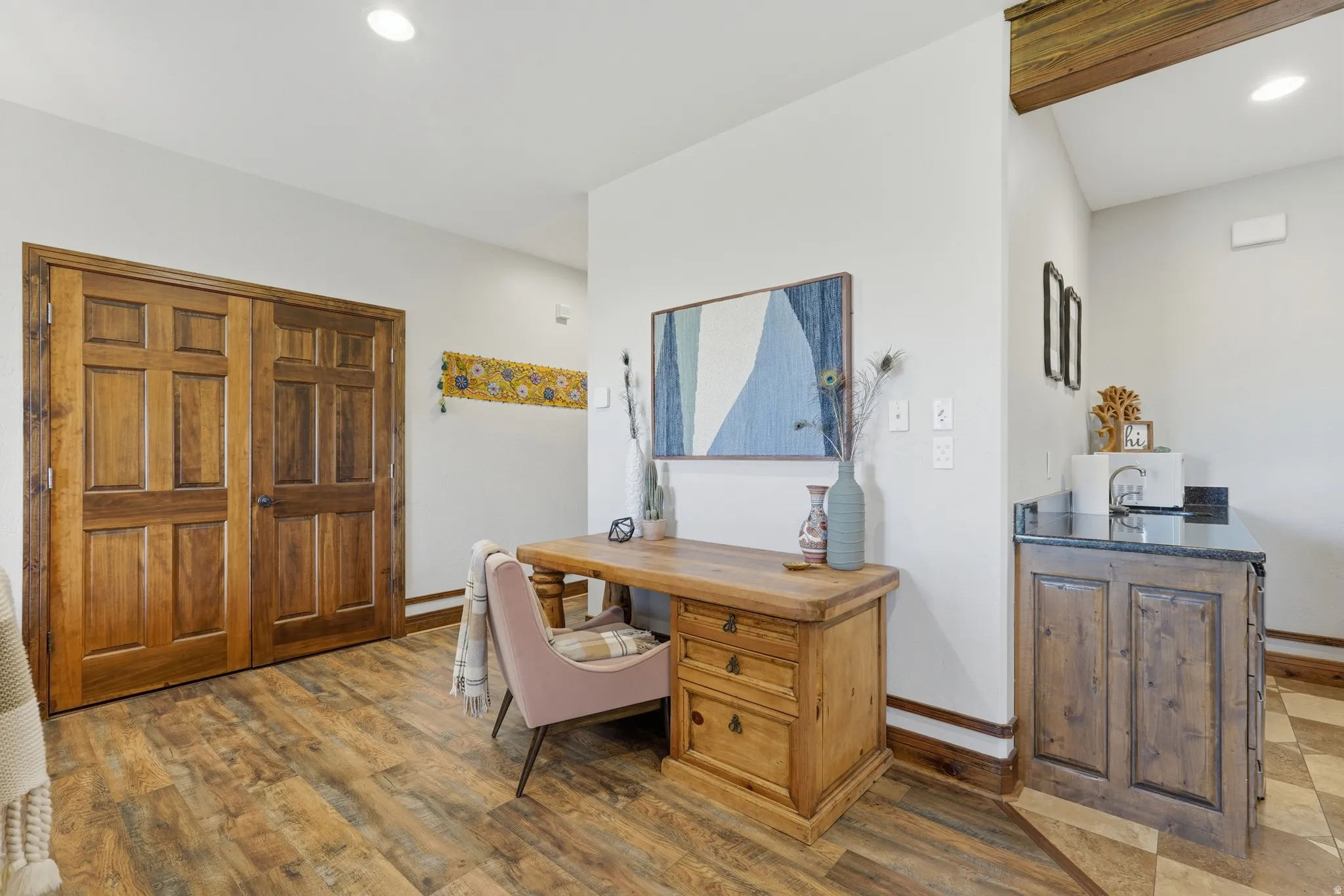 Rich wood-finish flooring provides a warm foundation for this versatile space featuring paneled wood doors, a built-in wet bar with a dark stone countertop, and recessed lighting
