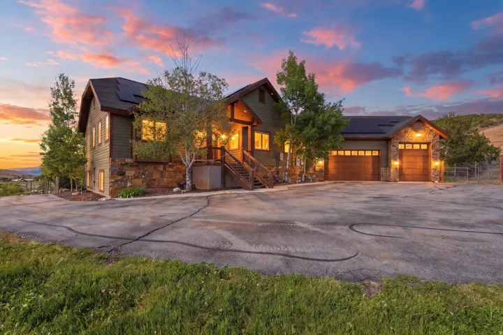 Exterior featuring dark green siding, natural stone accents, a multi-pane garage door, dark shingle roofing, and established trees