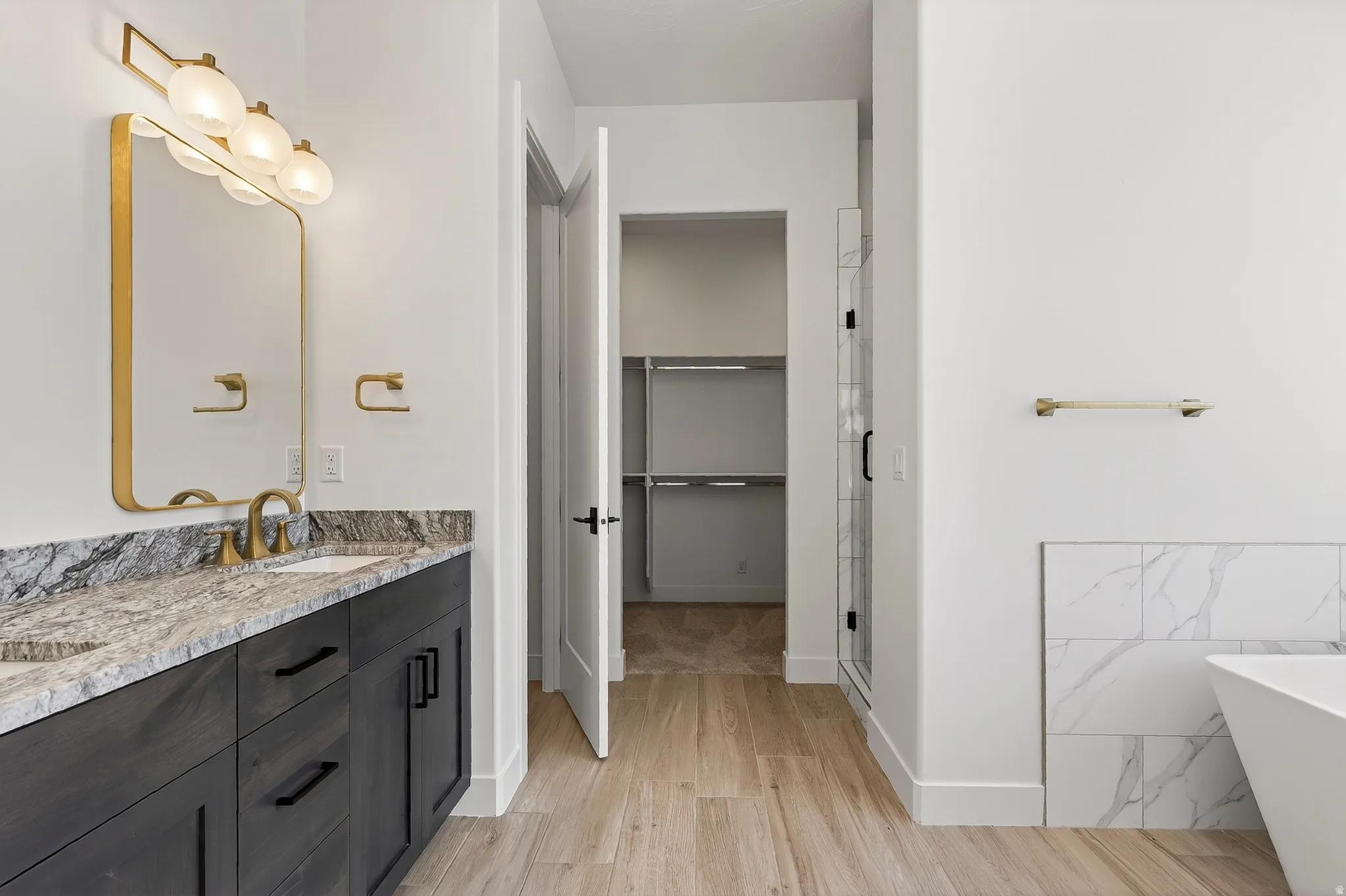 Bathroom featuring a double vanity with stone countertops, a freestanding tub, a glass-enclosed shower, wood-finish flooring, and a walk-in closet