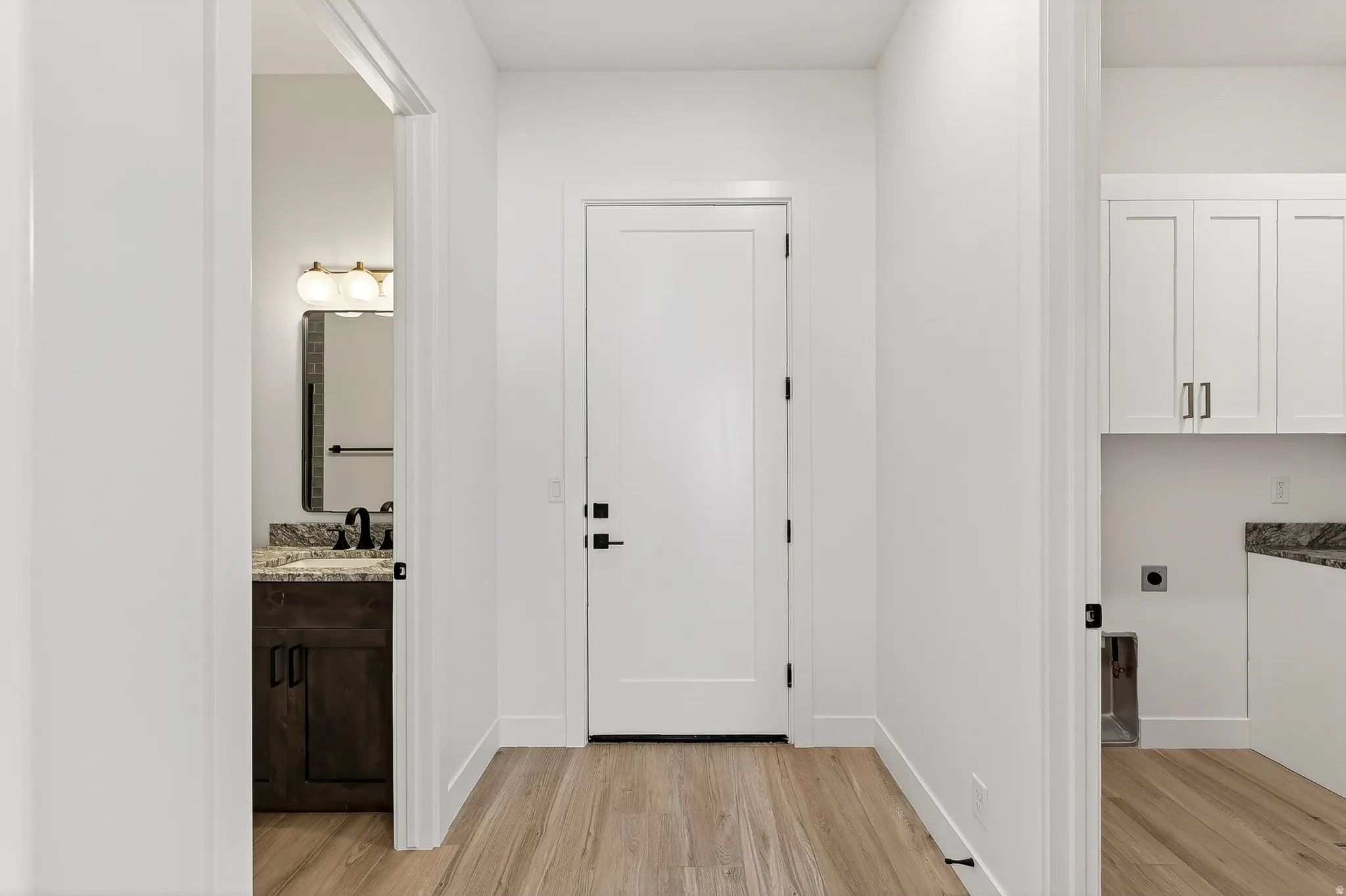 Hallway with wood-finish flooring, featuring a solid core door with matte black hardware, and views into a vanity area with a granite countertop and a utility area with white cabinetry